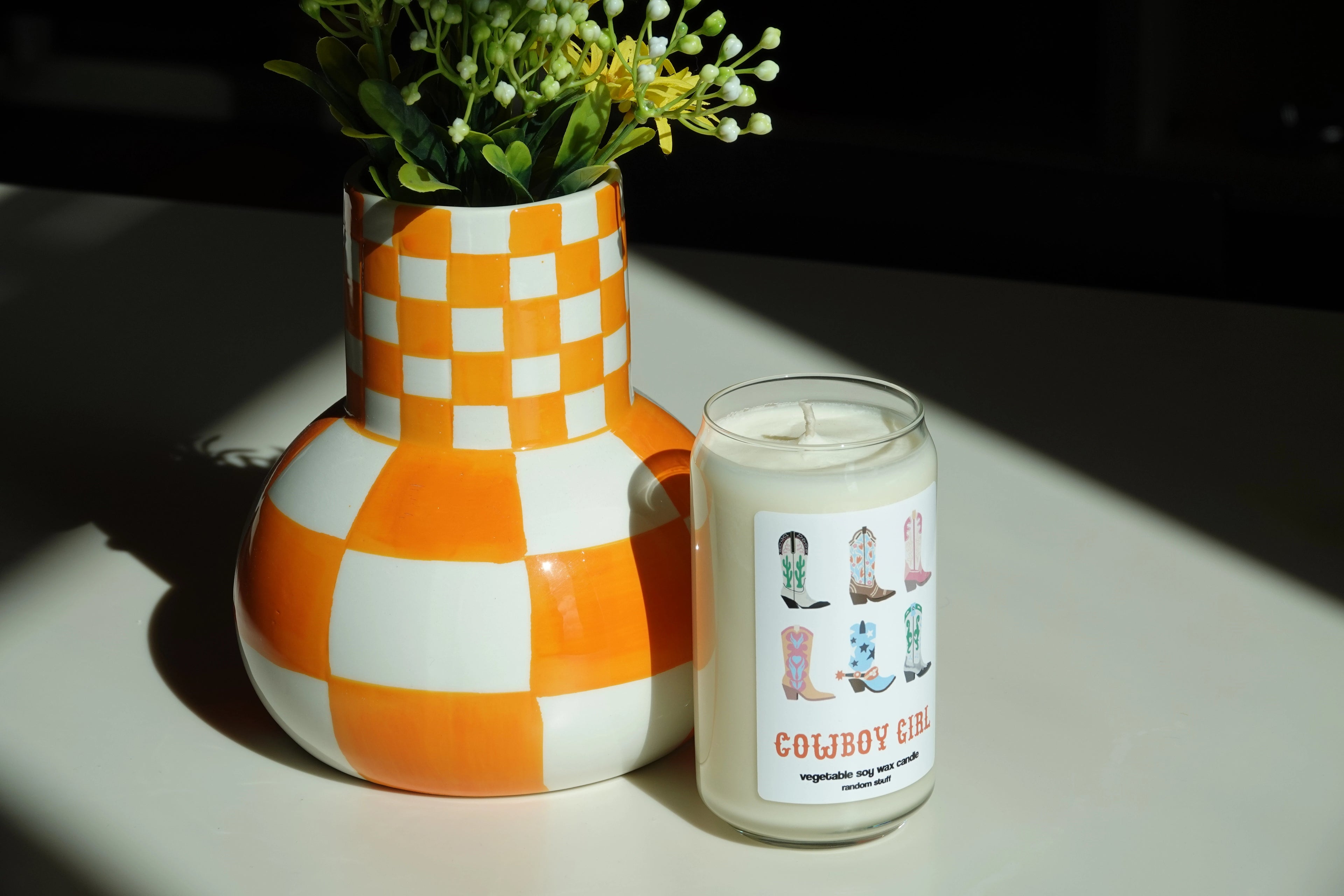 Handcrafted soy wax candle in a Cowboy Girl design, placed next to a checkered orange and white vase with greenery. Perfect for adding a touch of personality and fragrance to any space.